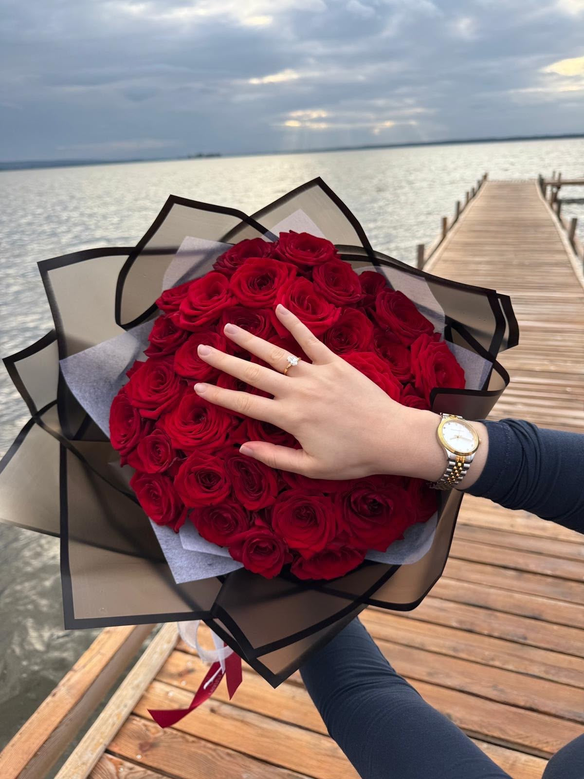 Hand mit Verlobungsring auf einem Strauß roter Rosen am Wasser